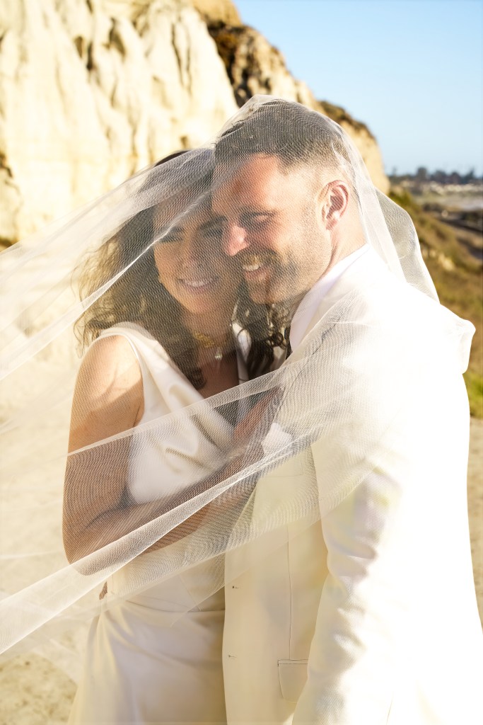 A joyful bride and groom embrace under a veil, with a scenic coastal background.